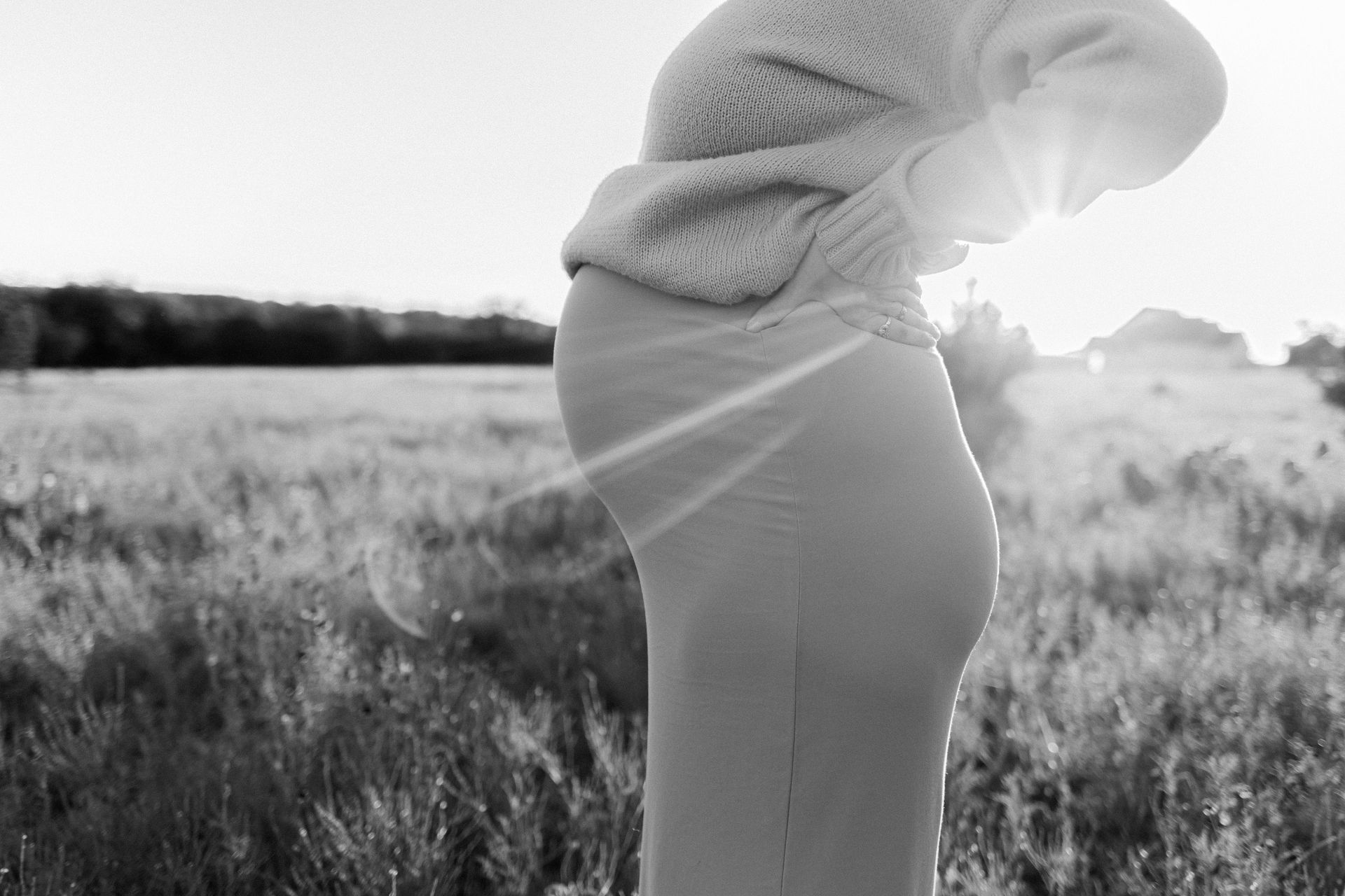 A pregnant woman is standing in a field with the sun shining on her belly.