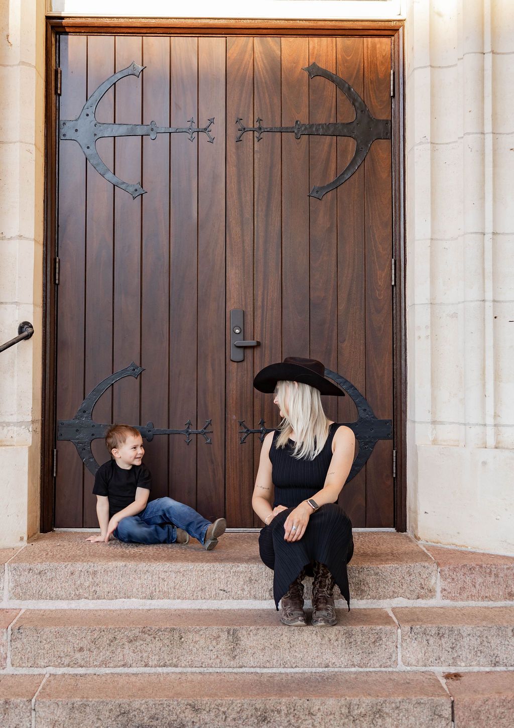 A woman and a child are sitting on the steps of a building in front of a wooden door.