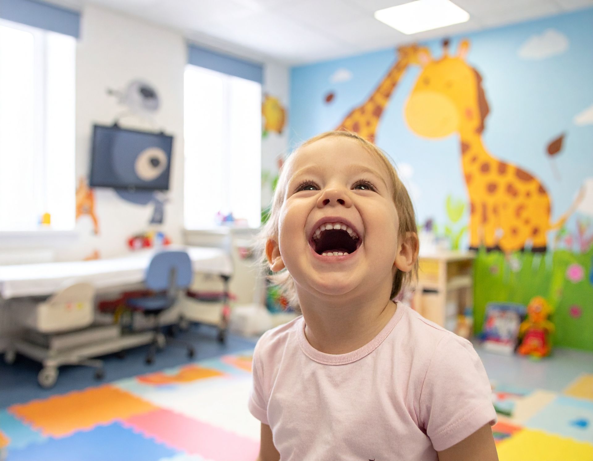 Smiling child in a colorful pediatric playroom with a giraffe mural and play mats