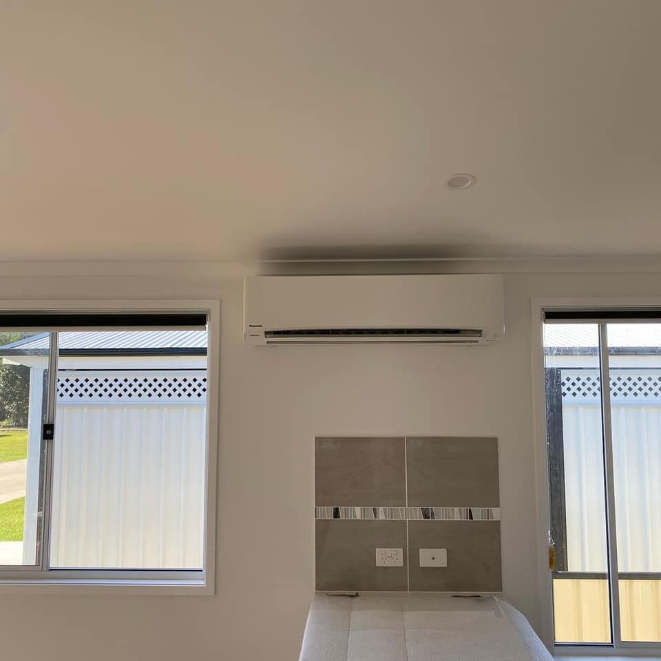White Air Conditioner on a Wall Above a Bed, Flanked by Windows — Airset Air Conditioning in Woolgoolga, NSW