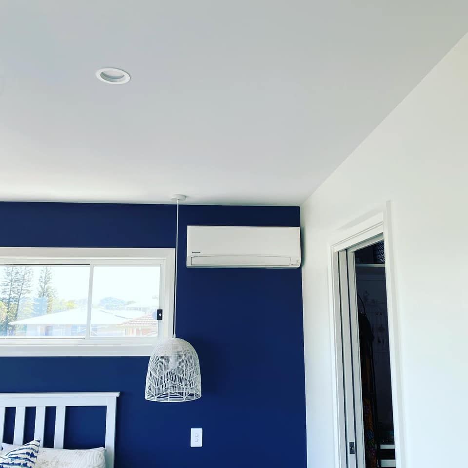 Bedroom corner with navy accent wall, white window and bed, wall-mounted AC, and hanging lamp — Airset Air Conditioning in Macksville, NSW