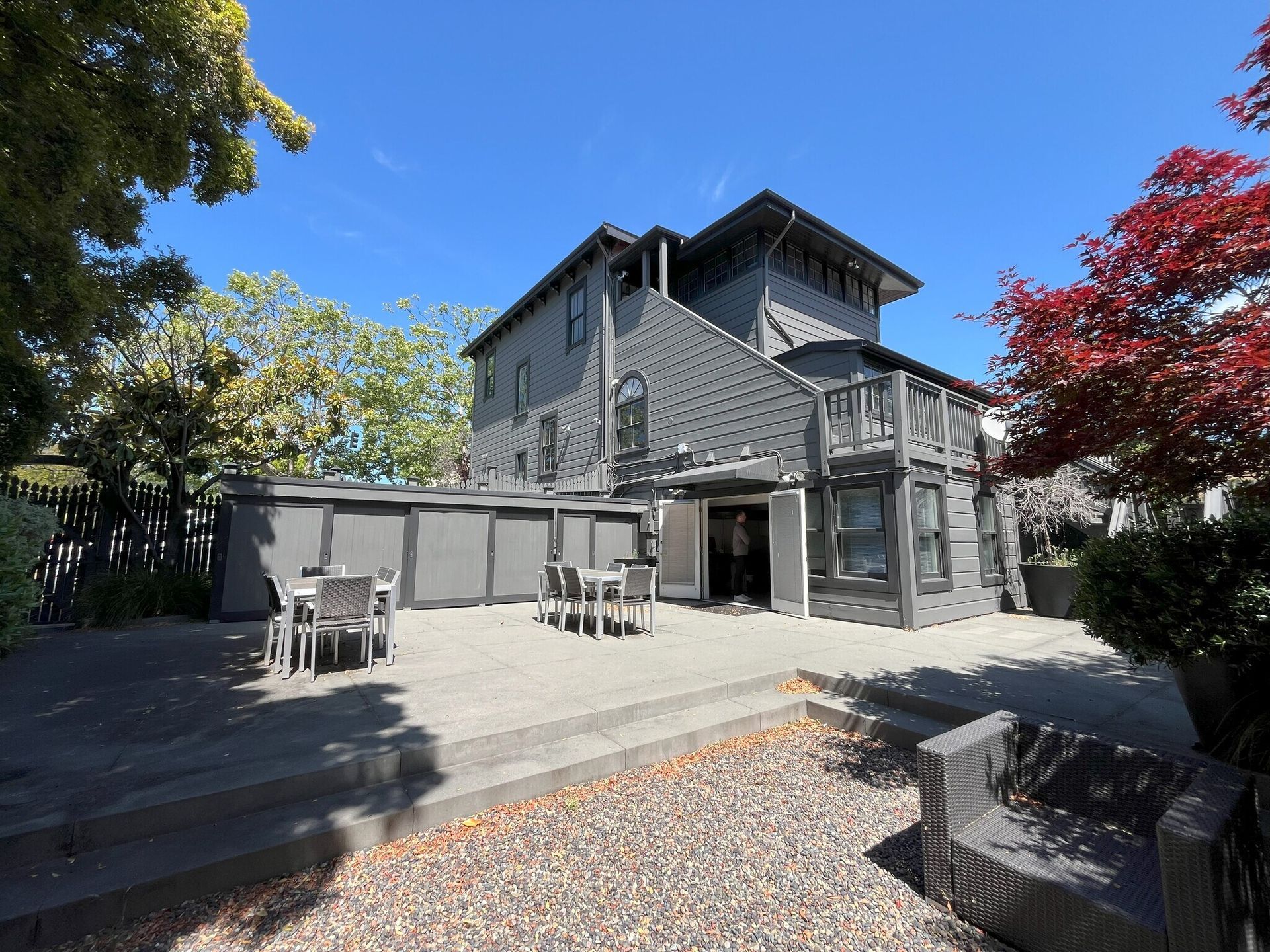 Gray two-story house with patio and tables, surrounded by trees under a blue sky.