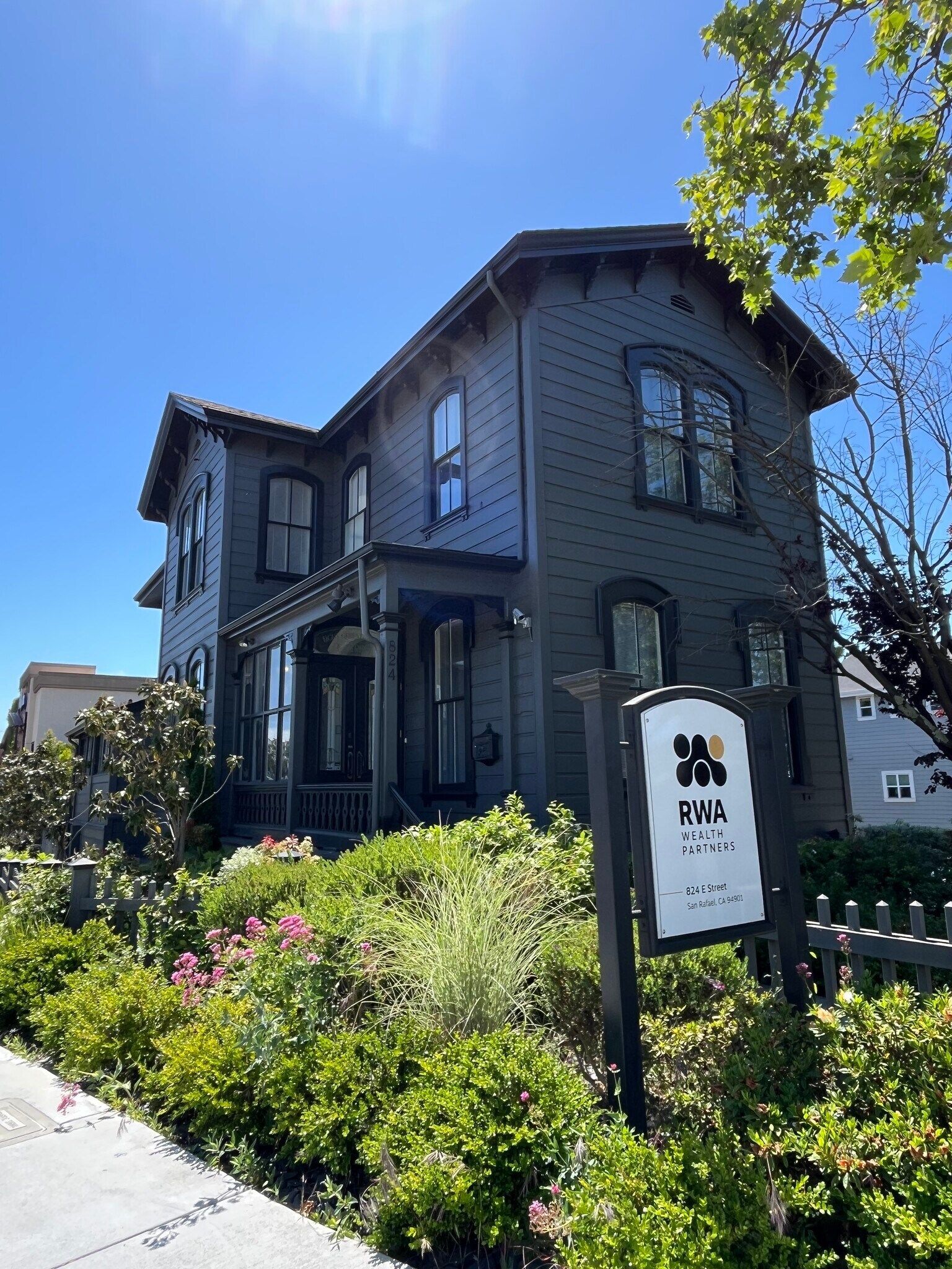 Dark grey Victorian house with a sign in front, surrounded by greenery.
