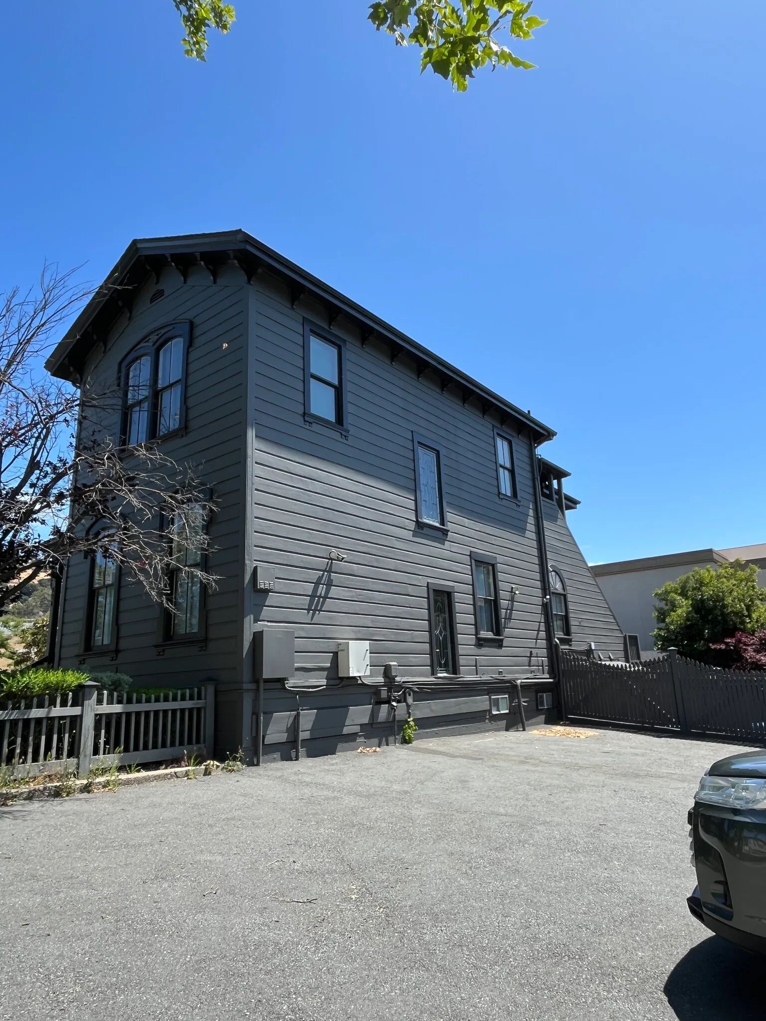 Two-story dark grey building with windows, a small fence, and a gravel parking area under a clear blue sky.