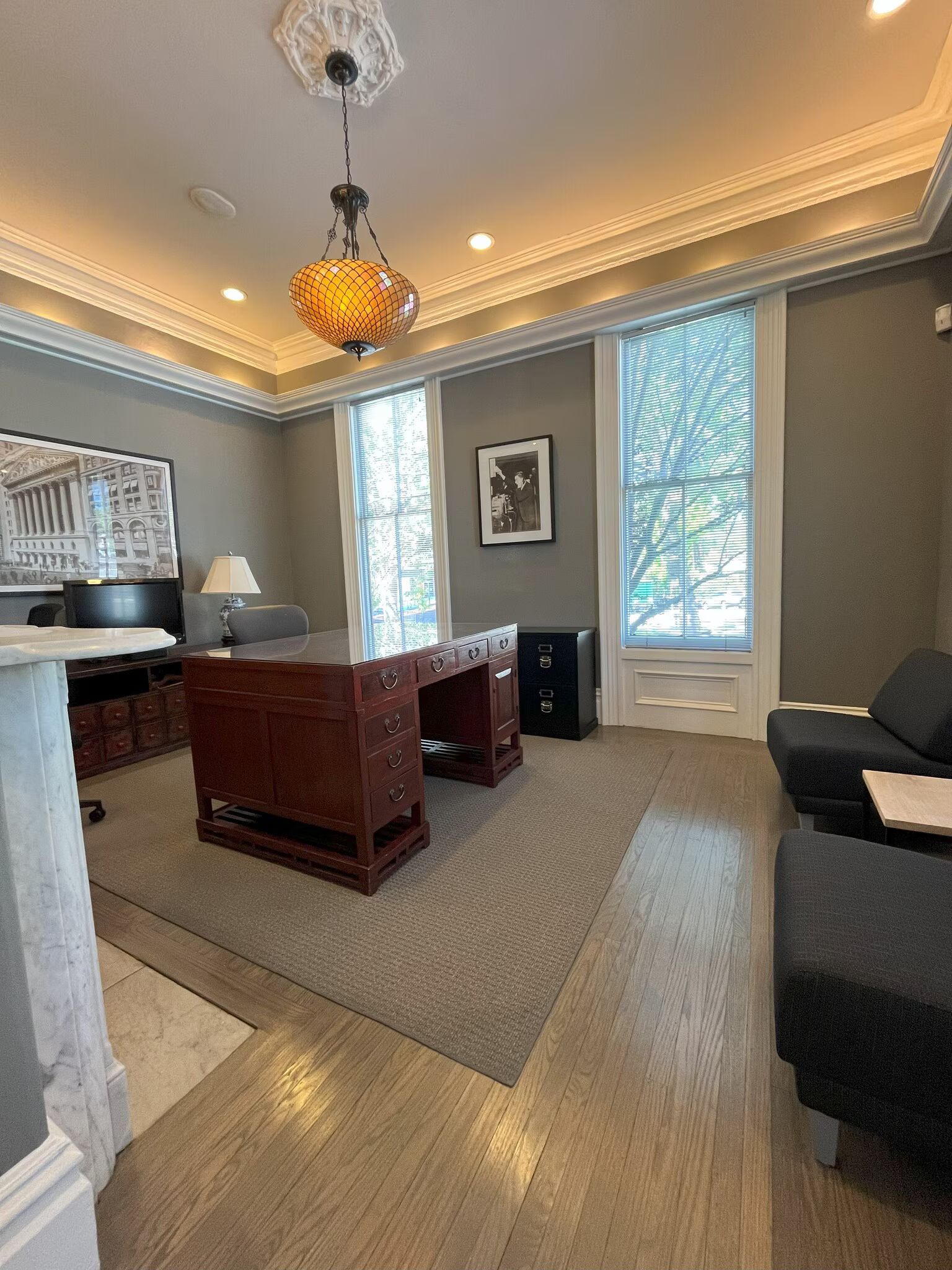 Office with wood desk, sofa, large windows, chandelier, and a framed art piece.