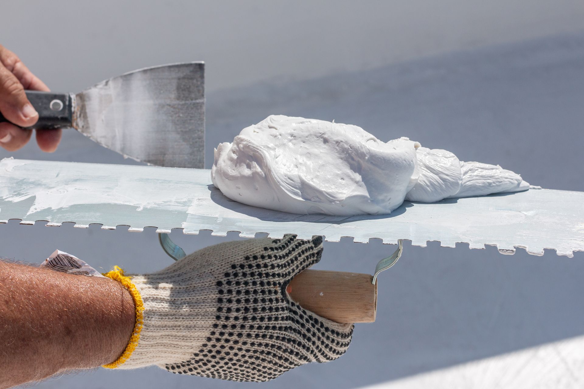 Person using a trowel to apply white compound to a surface with a notched spreader.
