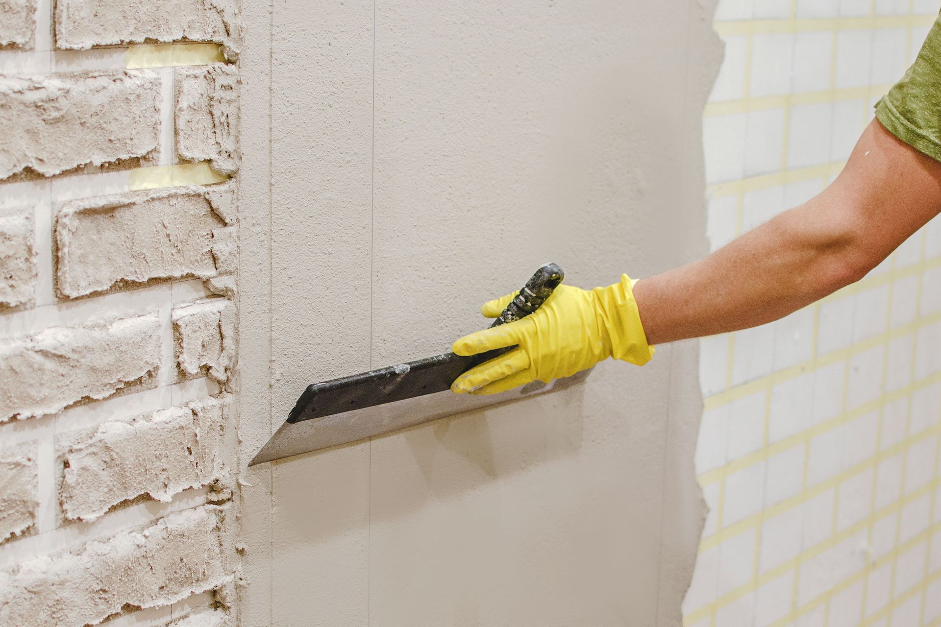 Person wearing yellow gloves smoothing plaster on wall with a trowel.