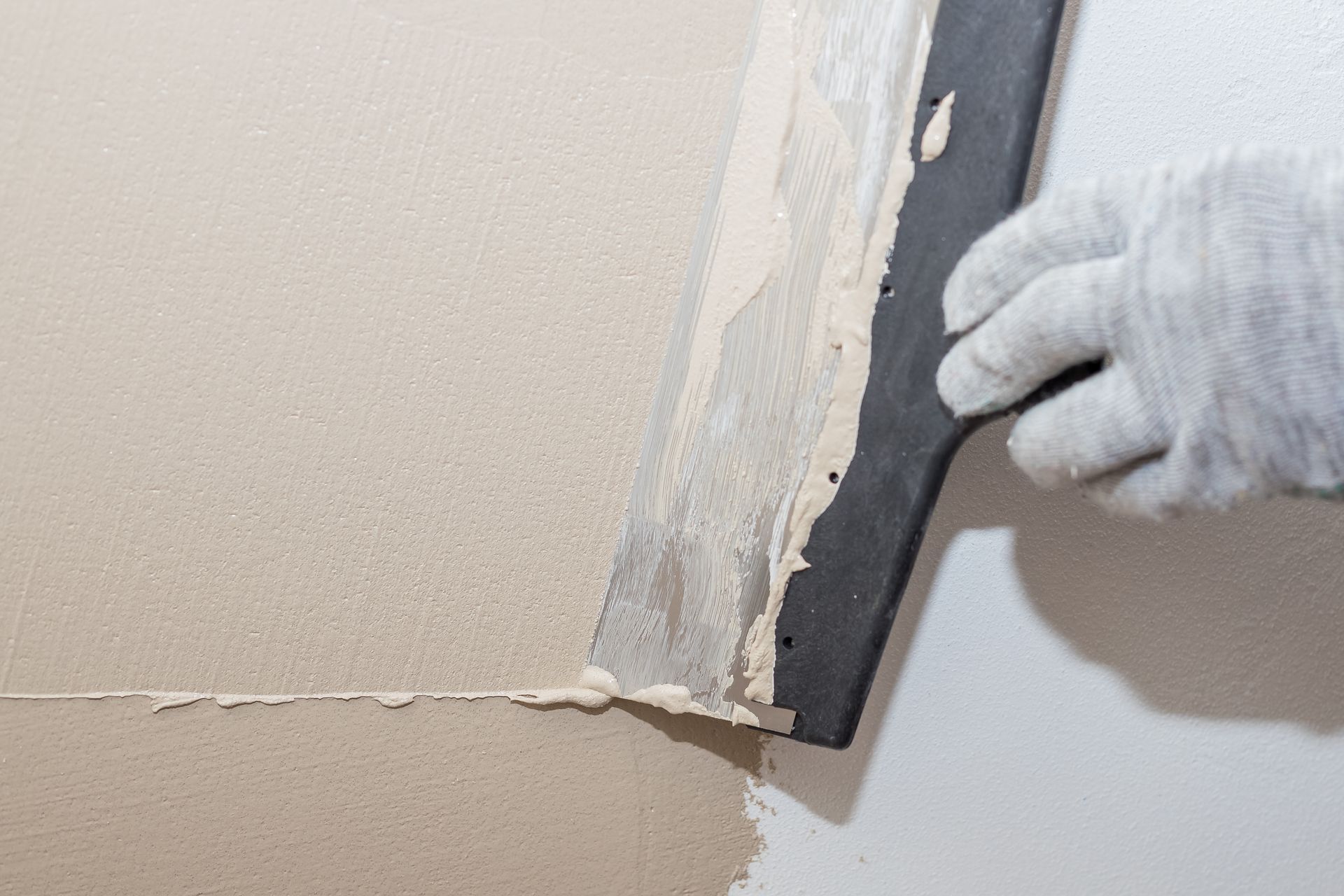 A gloved hand uses a putty knife to spread drywall compound on a wall.