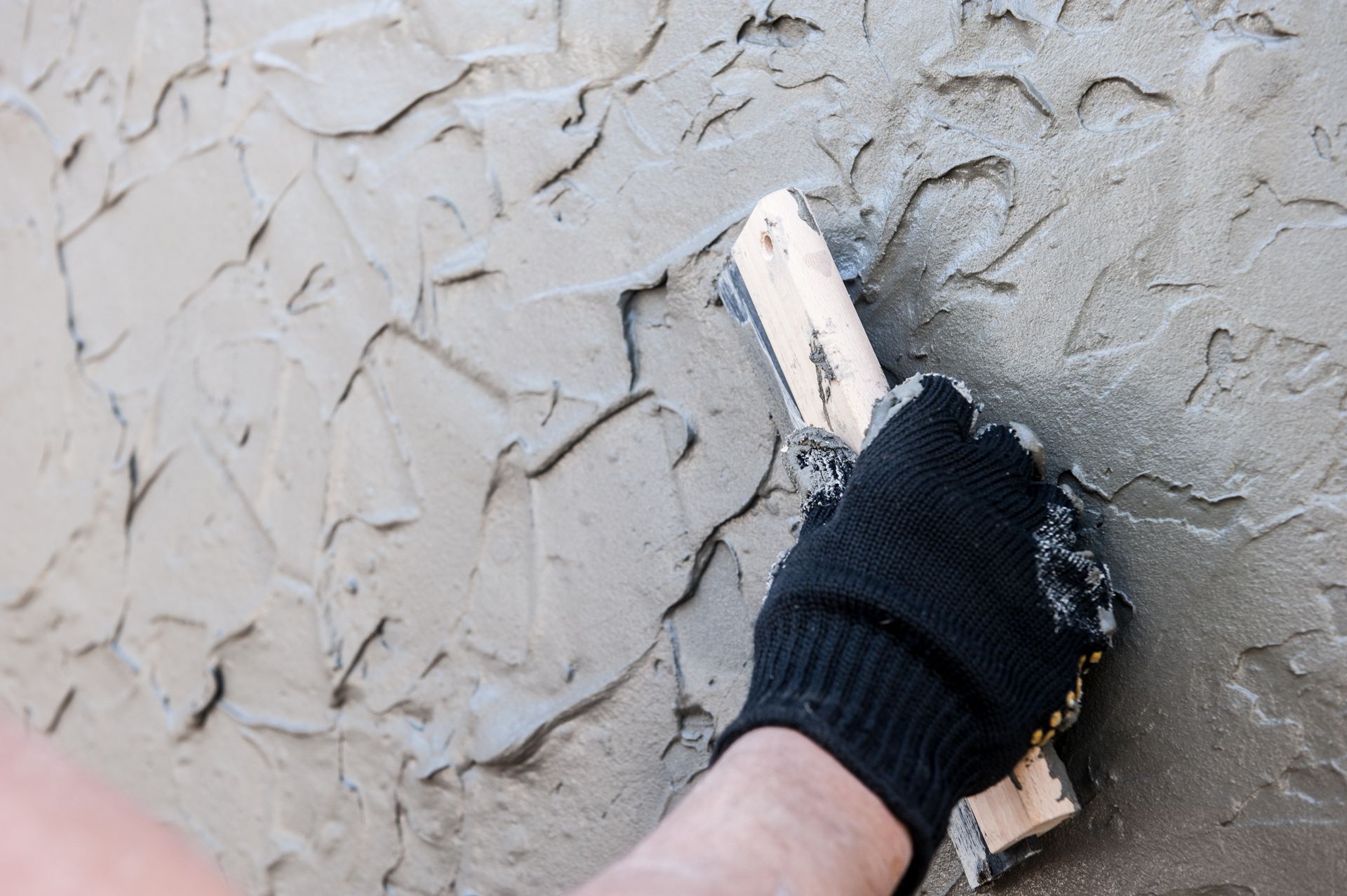 Hand wearing a black glove applying gray stucco to a wall with a wooden trowel.