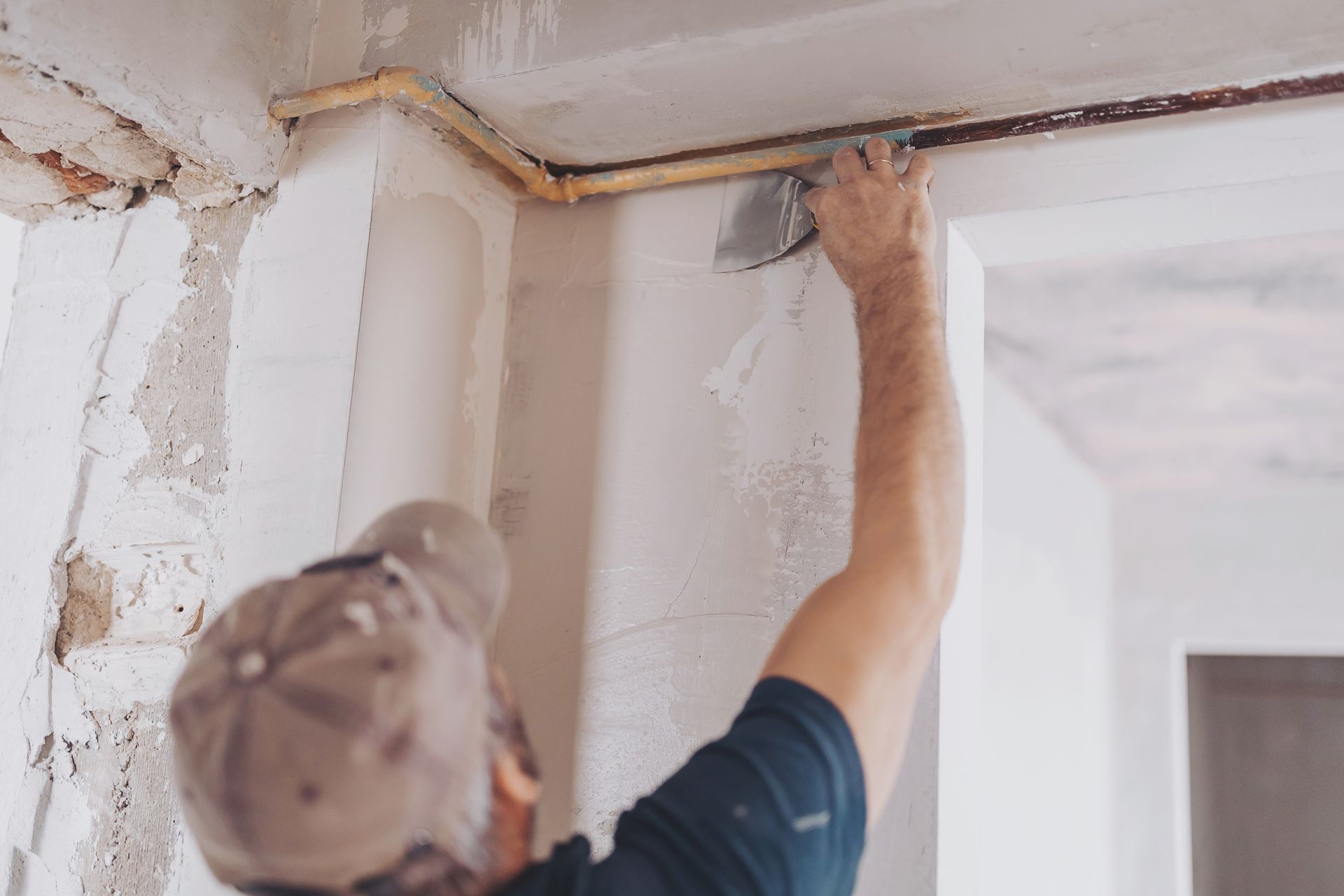Person applying plaster to a wall with a trowel, near exposed pipes.
