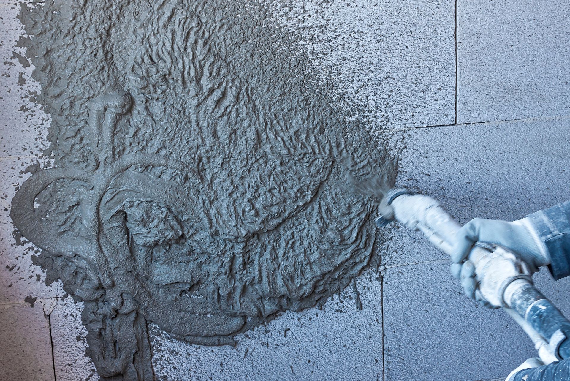 Person spraying concrete onto a gray brick wall with a nozzle.