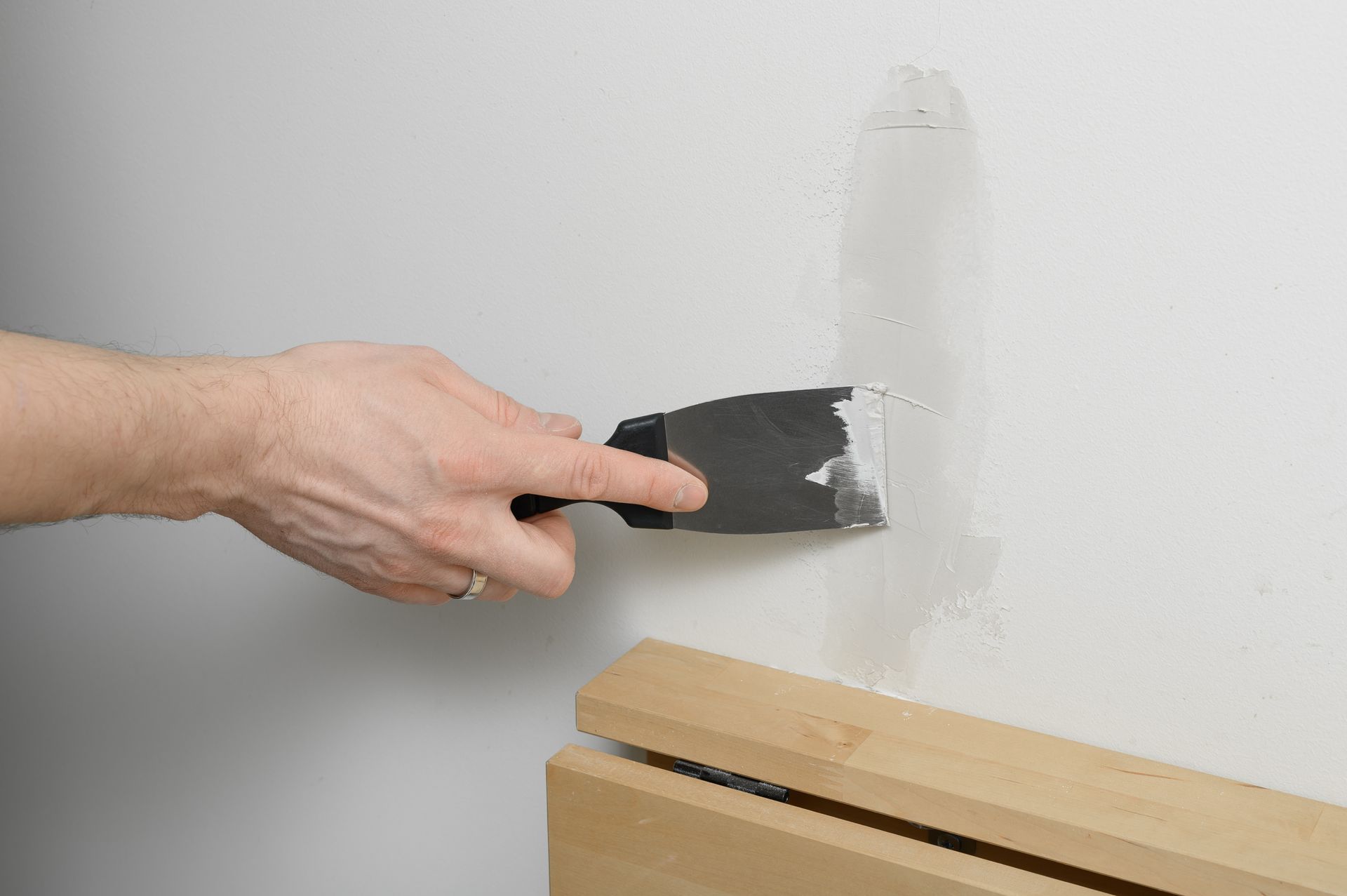 Hand applying spackle to a wall with a putty knife. Wooden trim piece visible below.