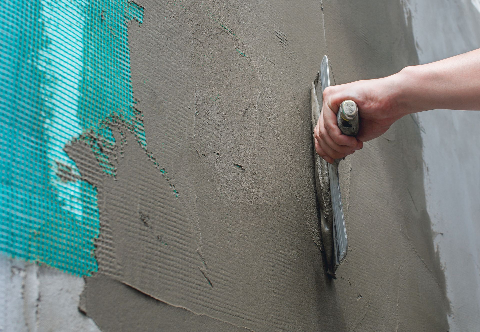 Person using a trowel to apply stucco over green mesh on a wall.