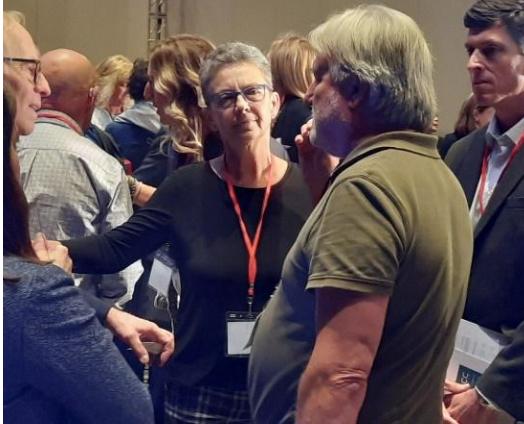 Woman in glasses talks to a man with gray hair in a crowd. People in background, conference setting.