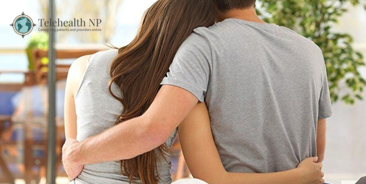 A couple embraces, arms around each other. The woman rests her head on the man's shoulder, indoor setting.