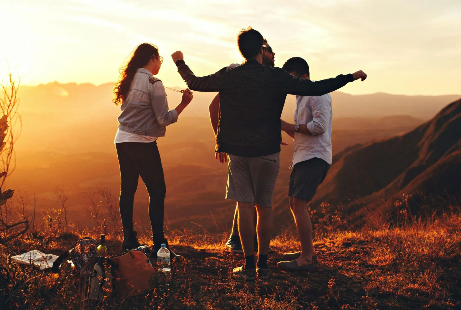 Friends celebrating on a mountain at sunset, arms raised. Golden light illuminates the group against a mountainous backdrop.