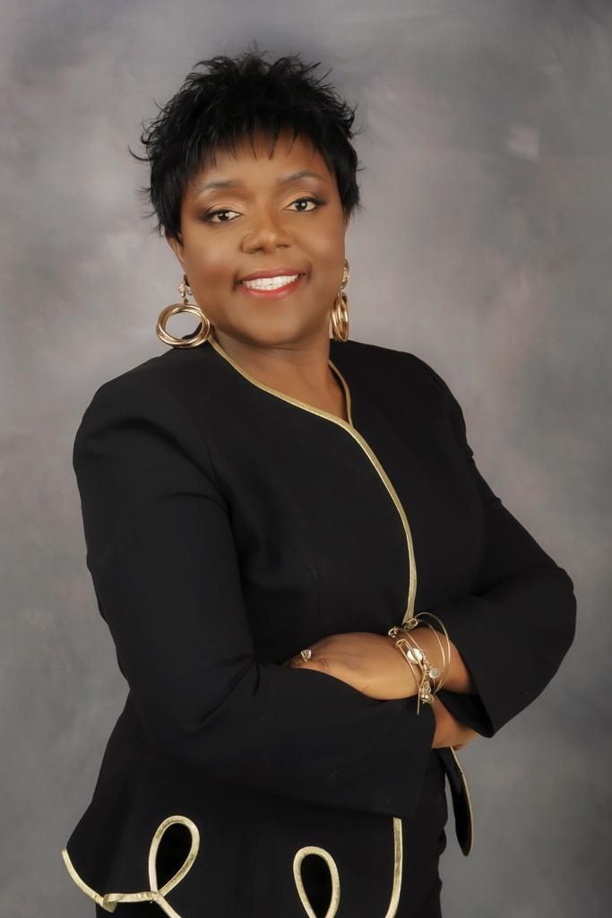 Woman with arms crossed, wearing black suit with gold trim, smiling.