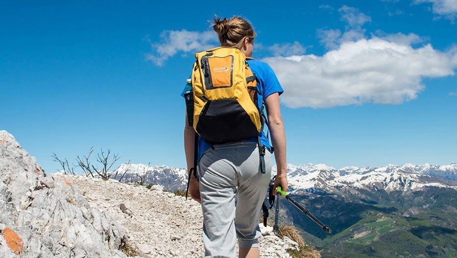 Woman hiking on a mountain ridge, wearing a yellow backpack, with snowy mountain range in background.