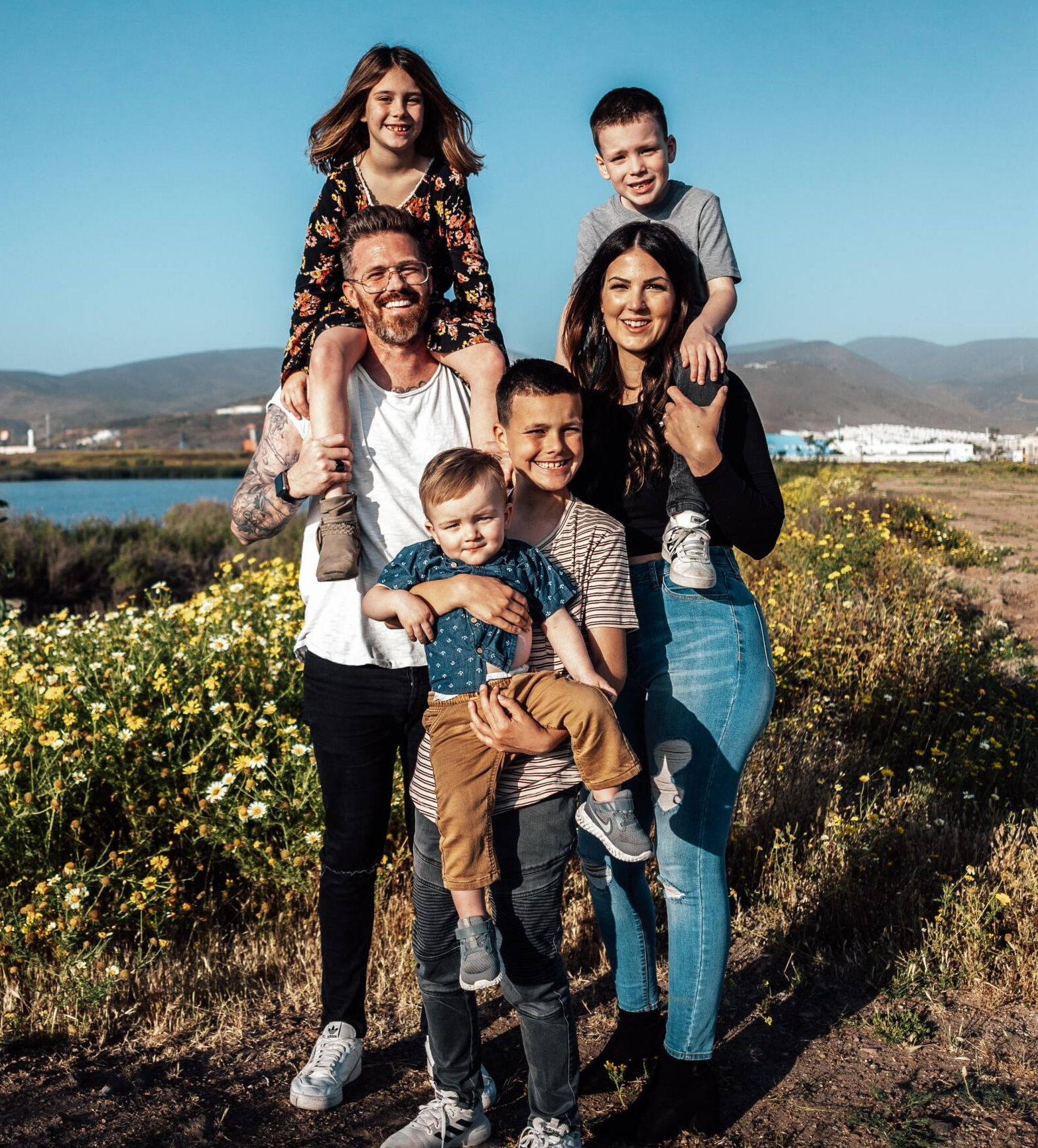 Family of seven poses outdoors, children on parents' shoulders, sunny day.