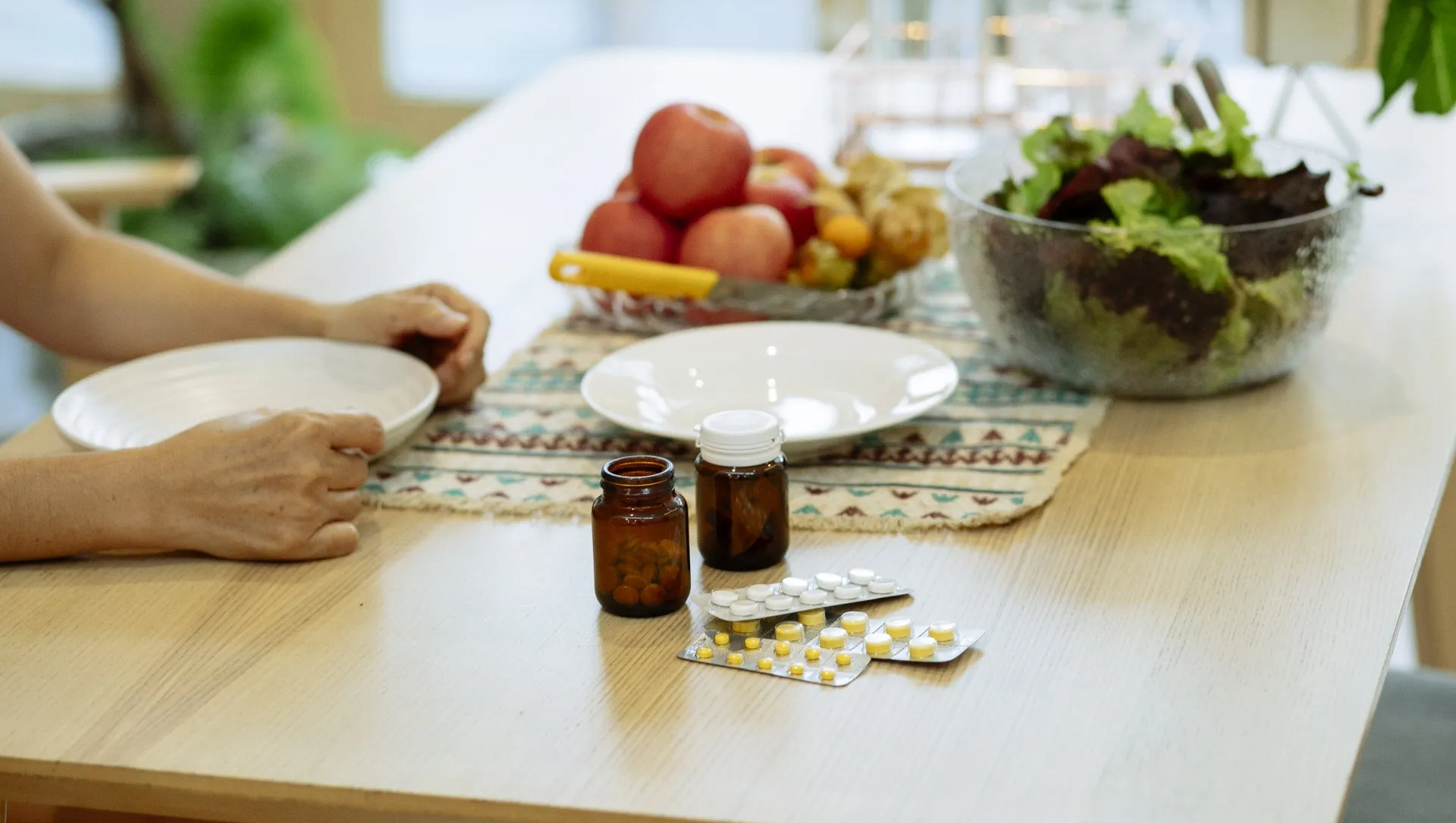Person at a table with pills, plates, fruit, and salad, likely preparing to take medication with a meal.