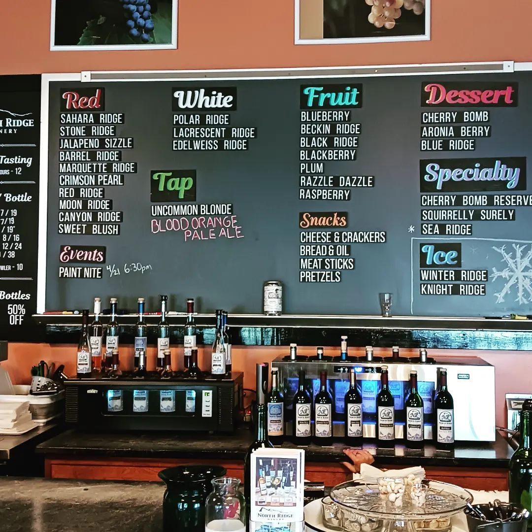 A menu board at a winery listing red, white, fruit, dessert, and specialty wines, alongside snacks and events.