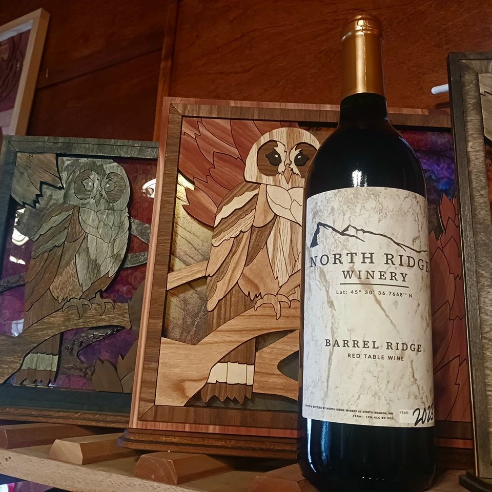 A bottle of North Ridge Winery red wine standing in front of two framed owl wood marquetry art pieces on a shelf.