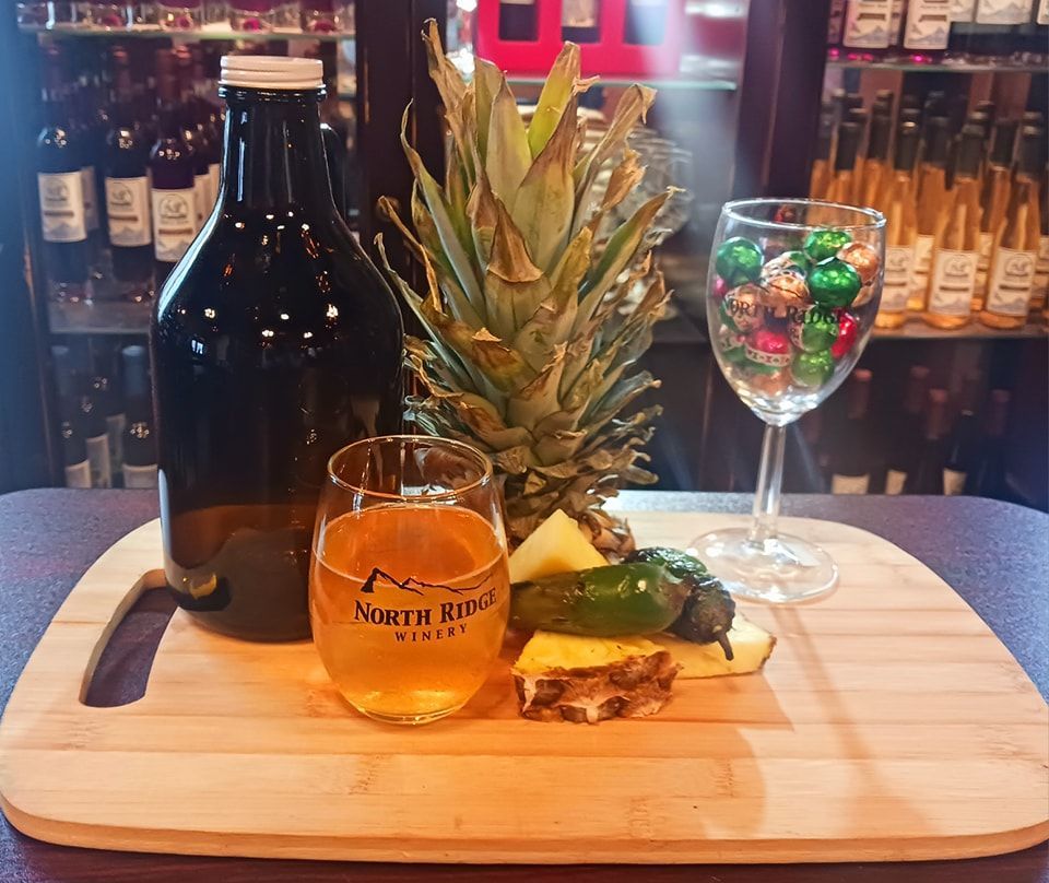 A wooden cutting board holds a brown bottle, a North Ridge Winery glass of cider, a pineapple, and colorful candies.