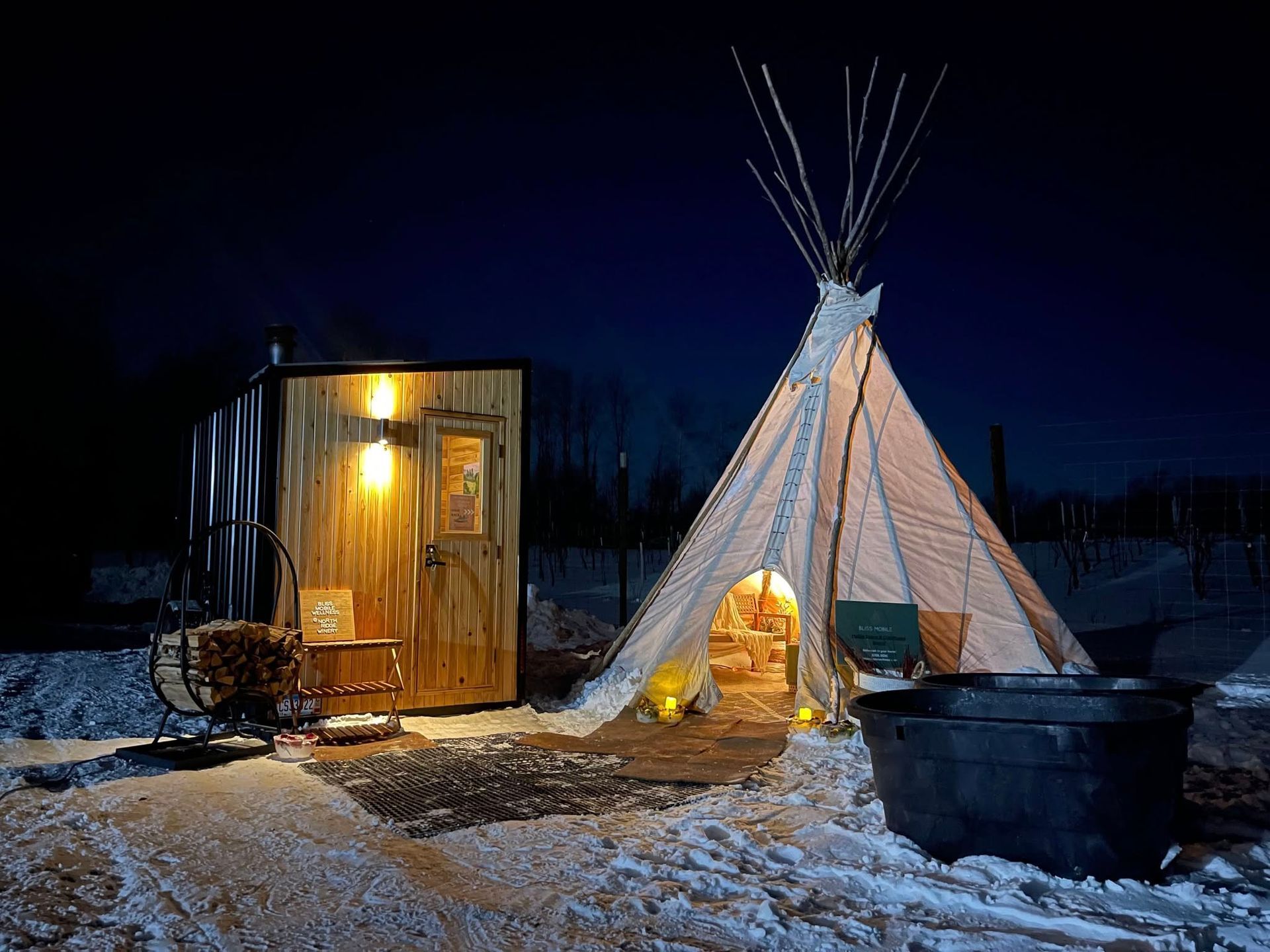 A brightly lit wooden sauna cabin next to a glowing teepee at night in a snowy landscape with a black soaking tub nearby.