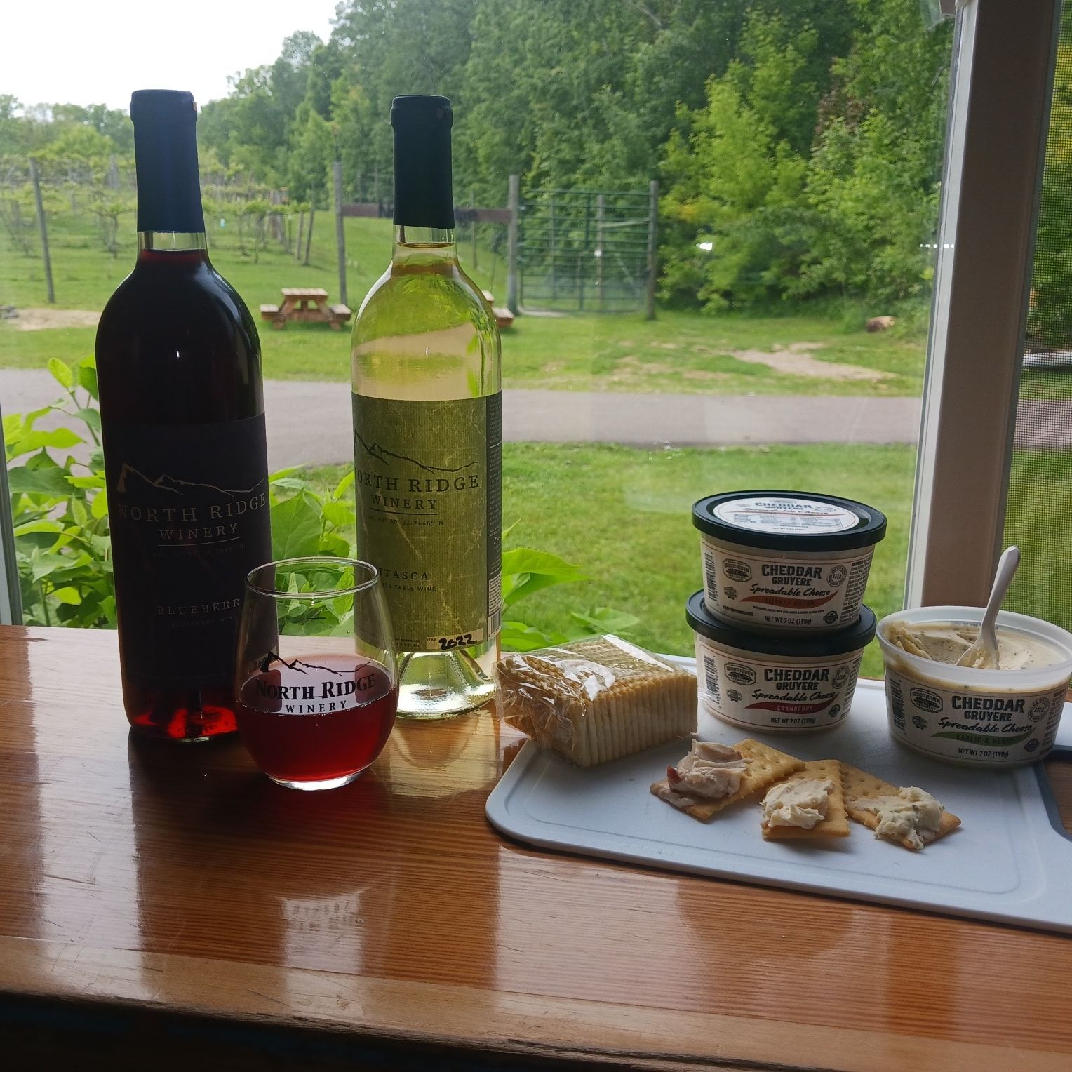 Two wine bottles, a glass of red wine, crackers, and tubs of dip are set on a table overlooking a lush green vineyard.