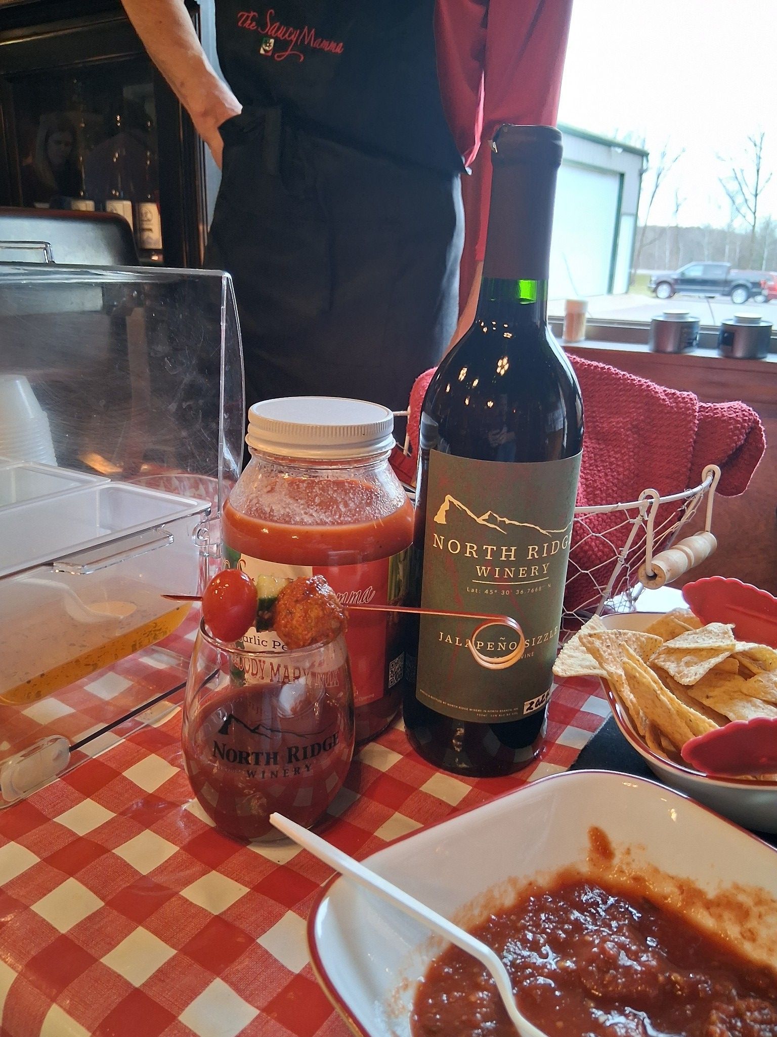 A table with a bottle of red wine, a jar of salsa, a cocktail with a garnish, and a bowl of dip on a red checkered cloth.