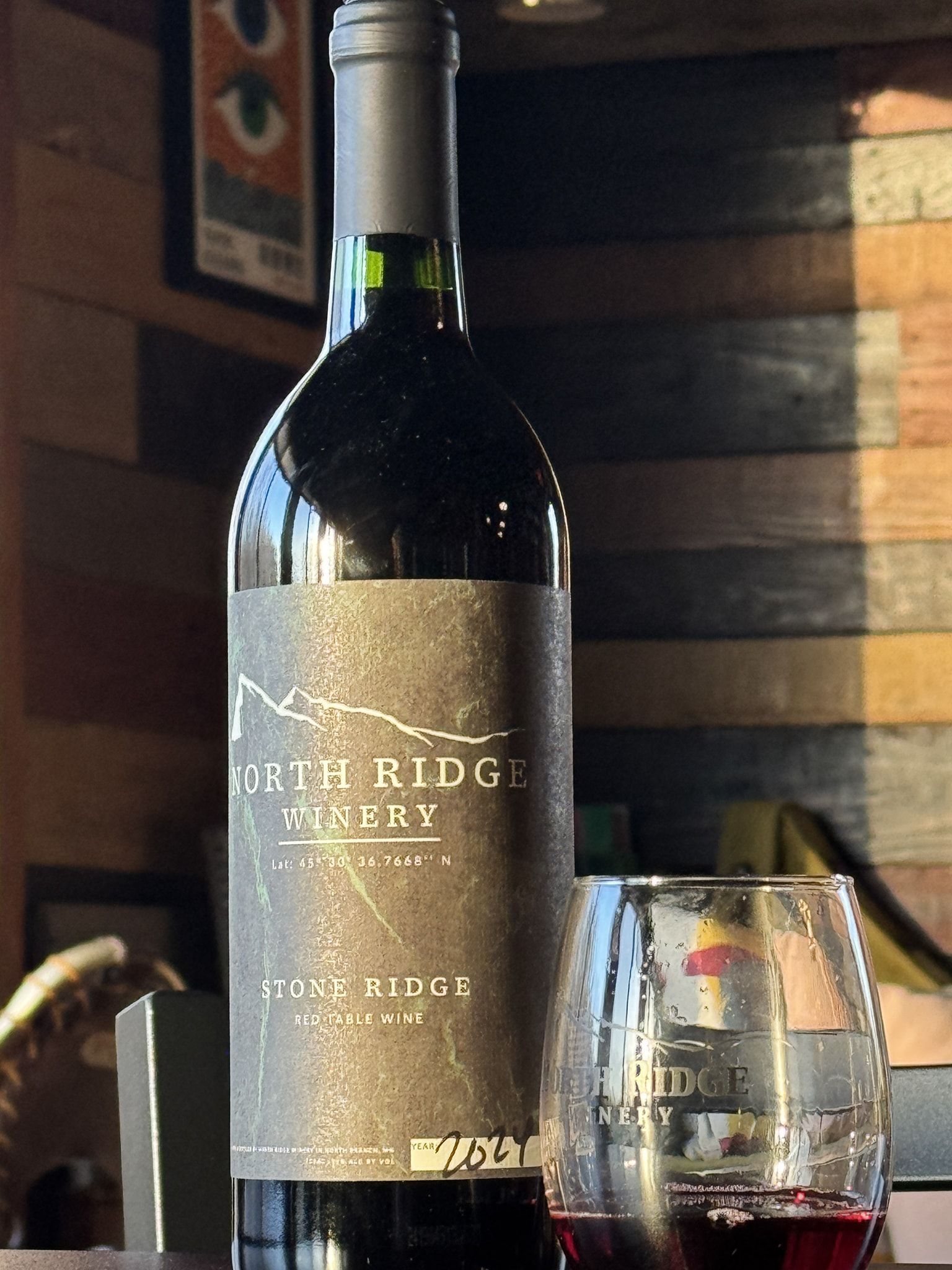 A bottle of North Ridge Winery red wine stands next to a glass of red wine on a table in front of a wood-paneled wall.