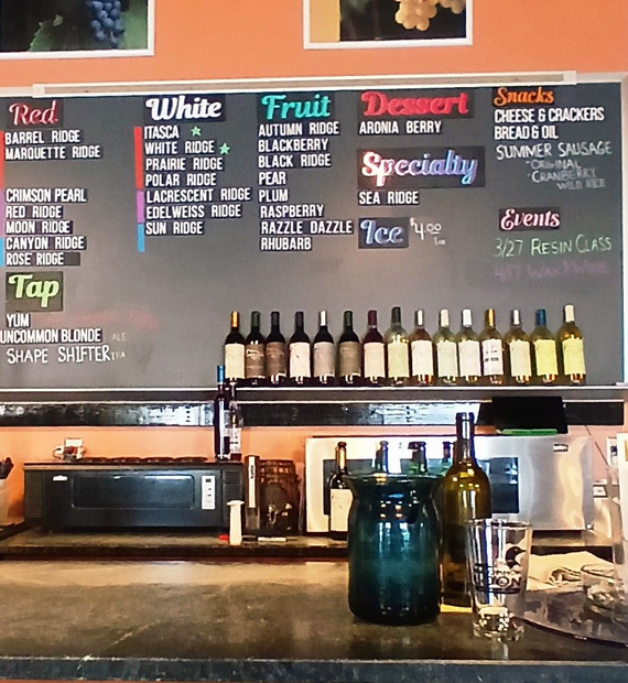 A menu board in a winery lists varieties of red, white, fruit, and dessert wines, plus specialty drinks, snacks, and events.