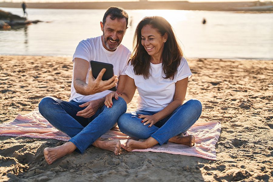 Couple on beach, smiling, looking at tablet screen together, sitting on a pink towel.