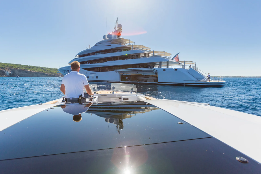 A man sits on the bow of a boat looking at a large yacht