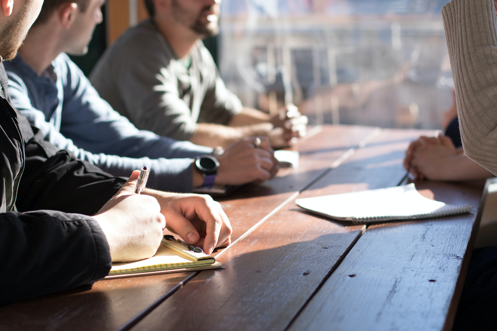 A group of men are sitting at a table writing in notebooks