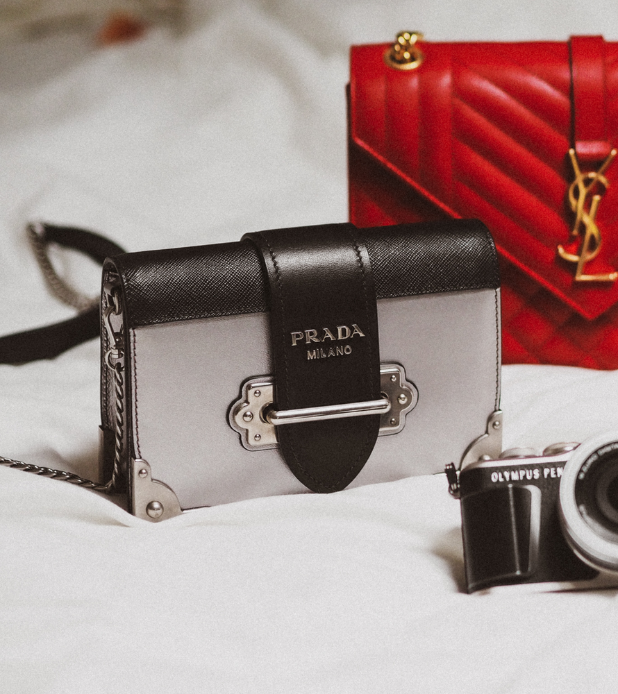 A prada purse sits on a bed next to a camera