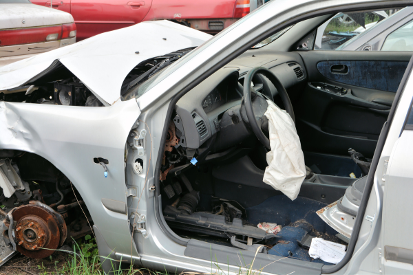 A wrecked silver sedan with a missing front wheel, crumpled hood, and deployed driver-side airbag.