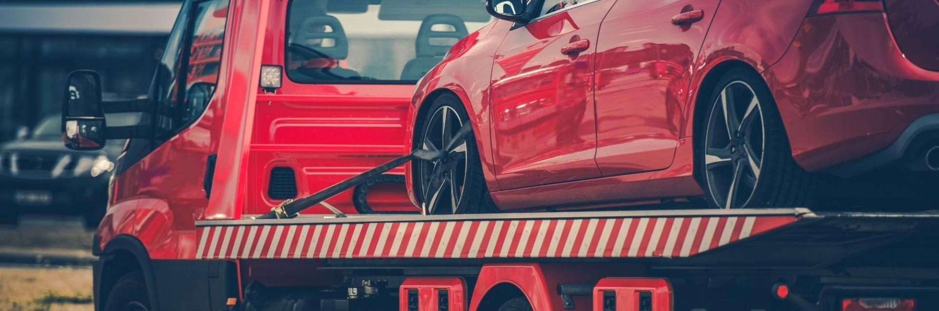A bright red flatbed tow truck transporting a matching red sedan, with a blurred dark car visible in the background.
