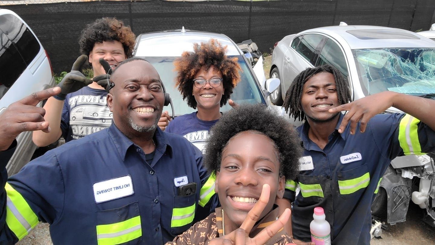 A group of five smiling individuals wearing blue work uniforms and safety gear pose for a selfie outdoors.