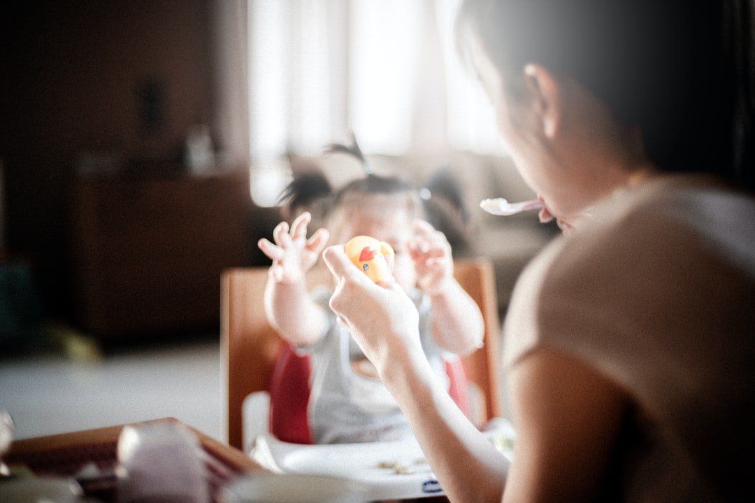 Person feeding a baby in a high chair at a table. Baby reaches for food with hands raised.