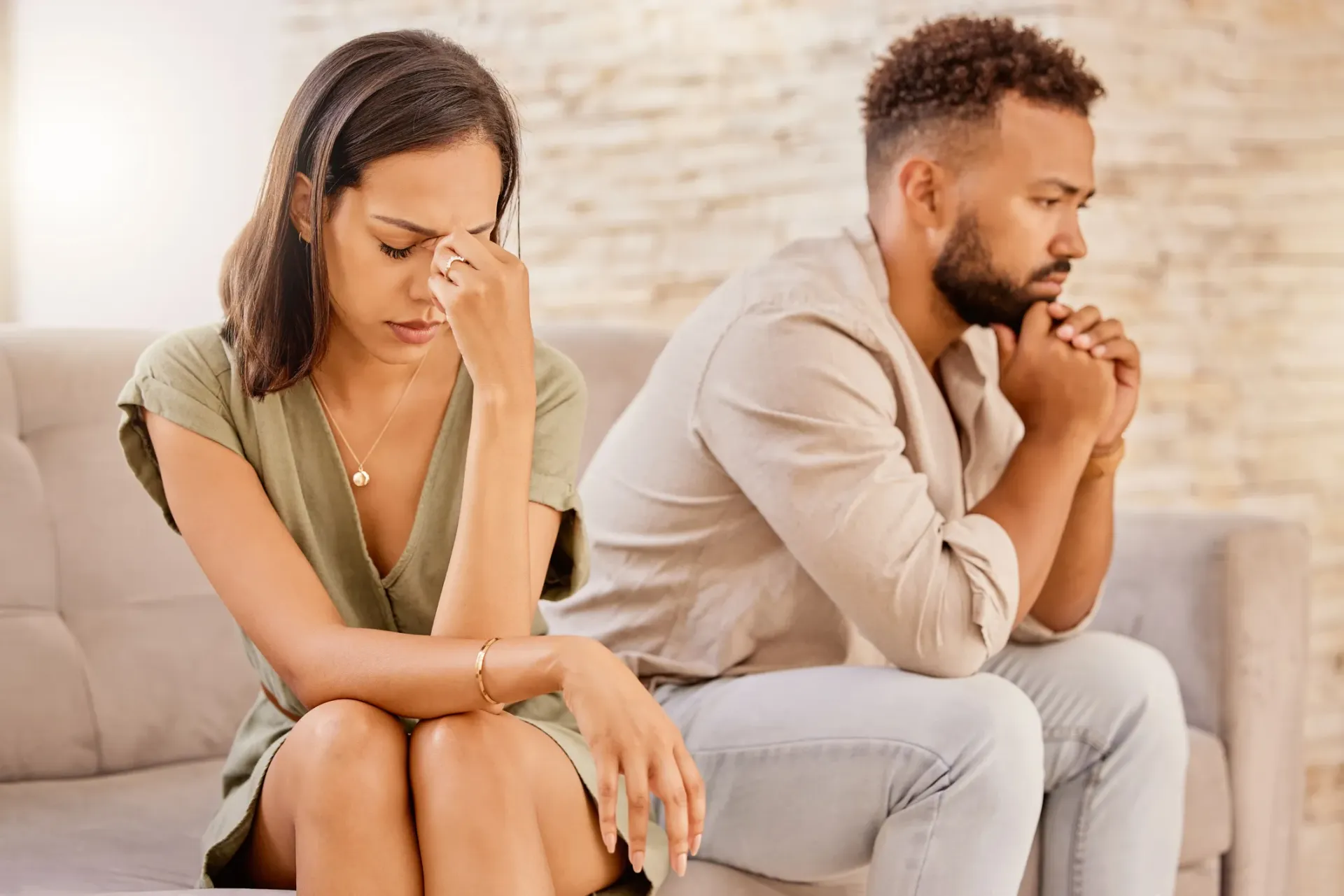 Couple sitting apart on a couch, looking sad. Woman holds her face, man looks down. Neutral color setting.