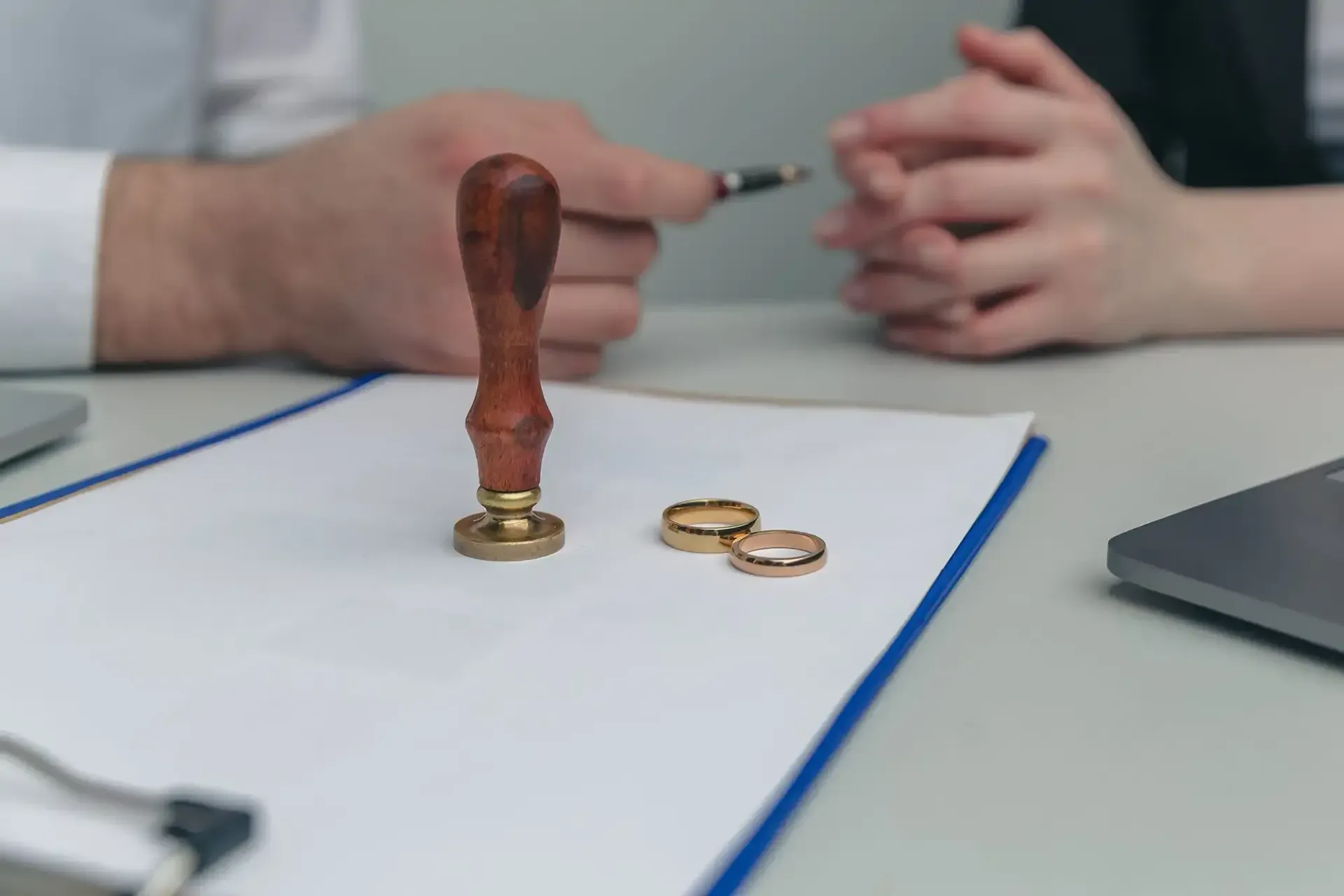 A wax seal stamp and two wedding rings sit on a document on a table between two people, suggesting a divorce proceeding.