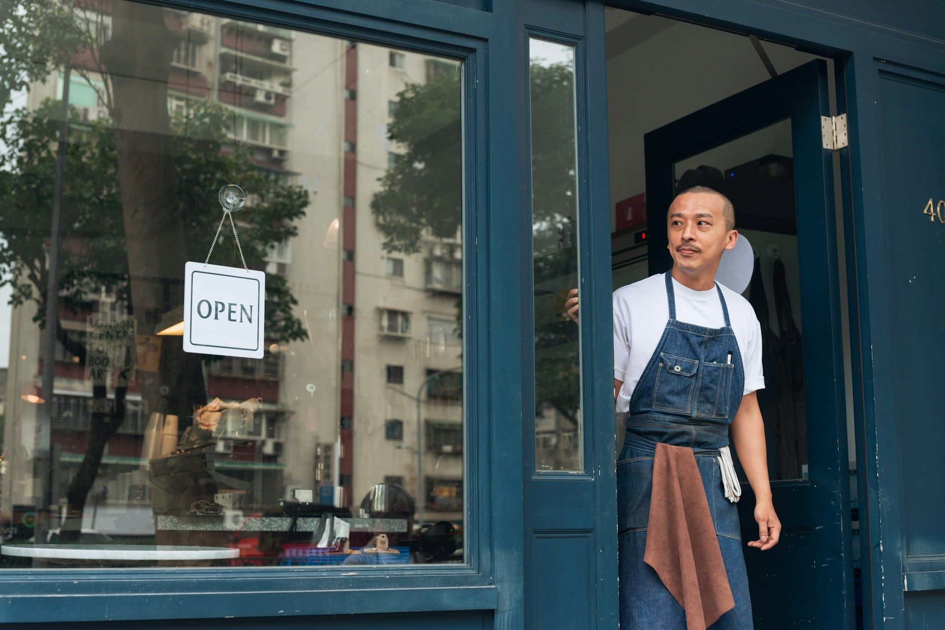 Shopkeeper standing in a blue doorway beside an “OPEN” sign, wearing an apron and smiling.