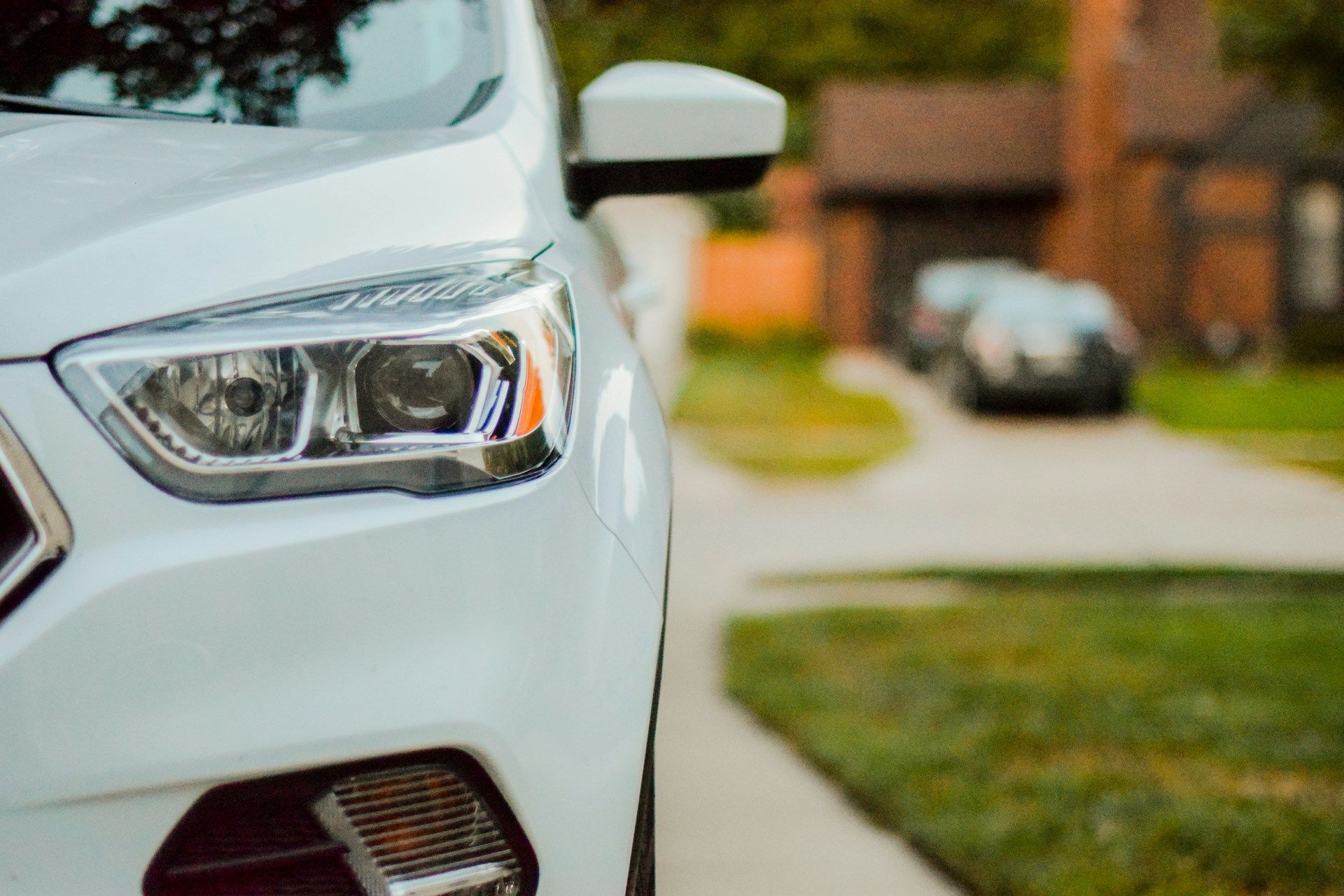 Front corner of a white car parked by a suburban sidewalk, with headlight and side mirror visible