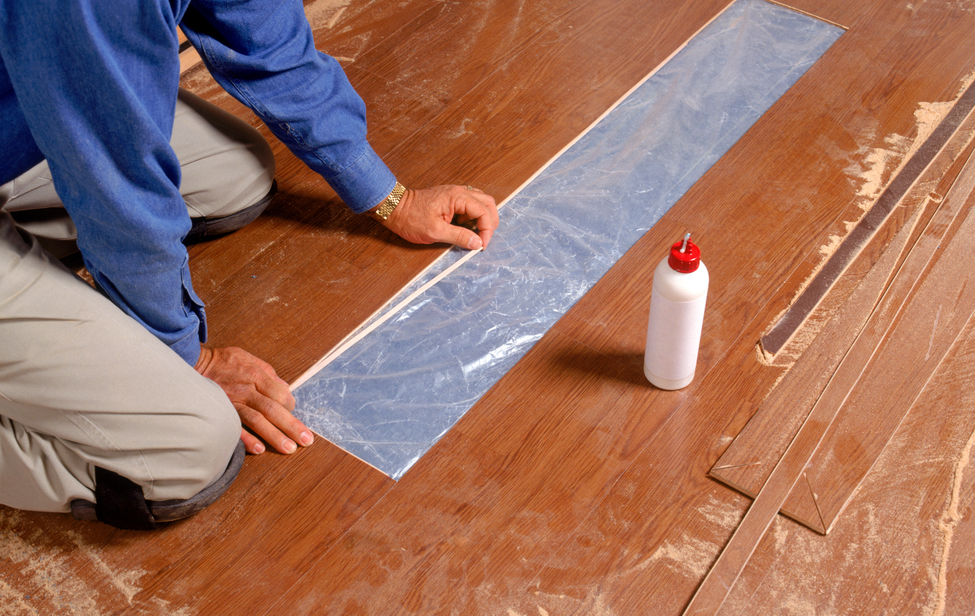 A man is kneeling down on a wooden floor next to a bottle of glue.