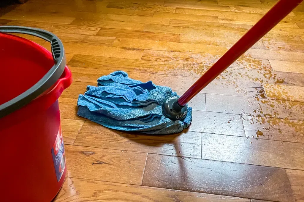 A mop is being used to clean a wooden floor next to a red bucket.