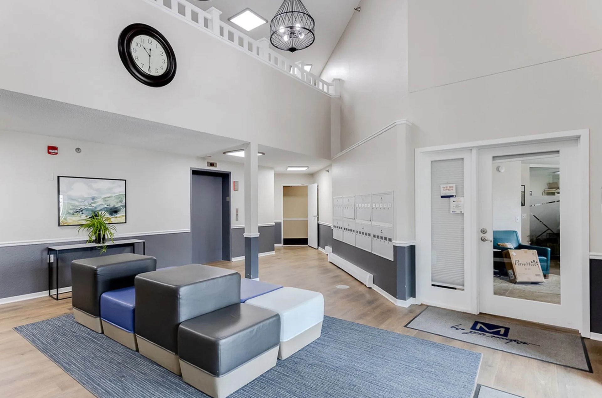 Lobby with seating, clock, and double doors. White walls with gray accents, wooden floor.