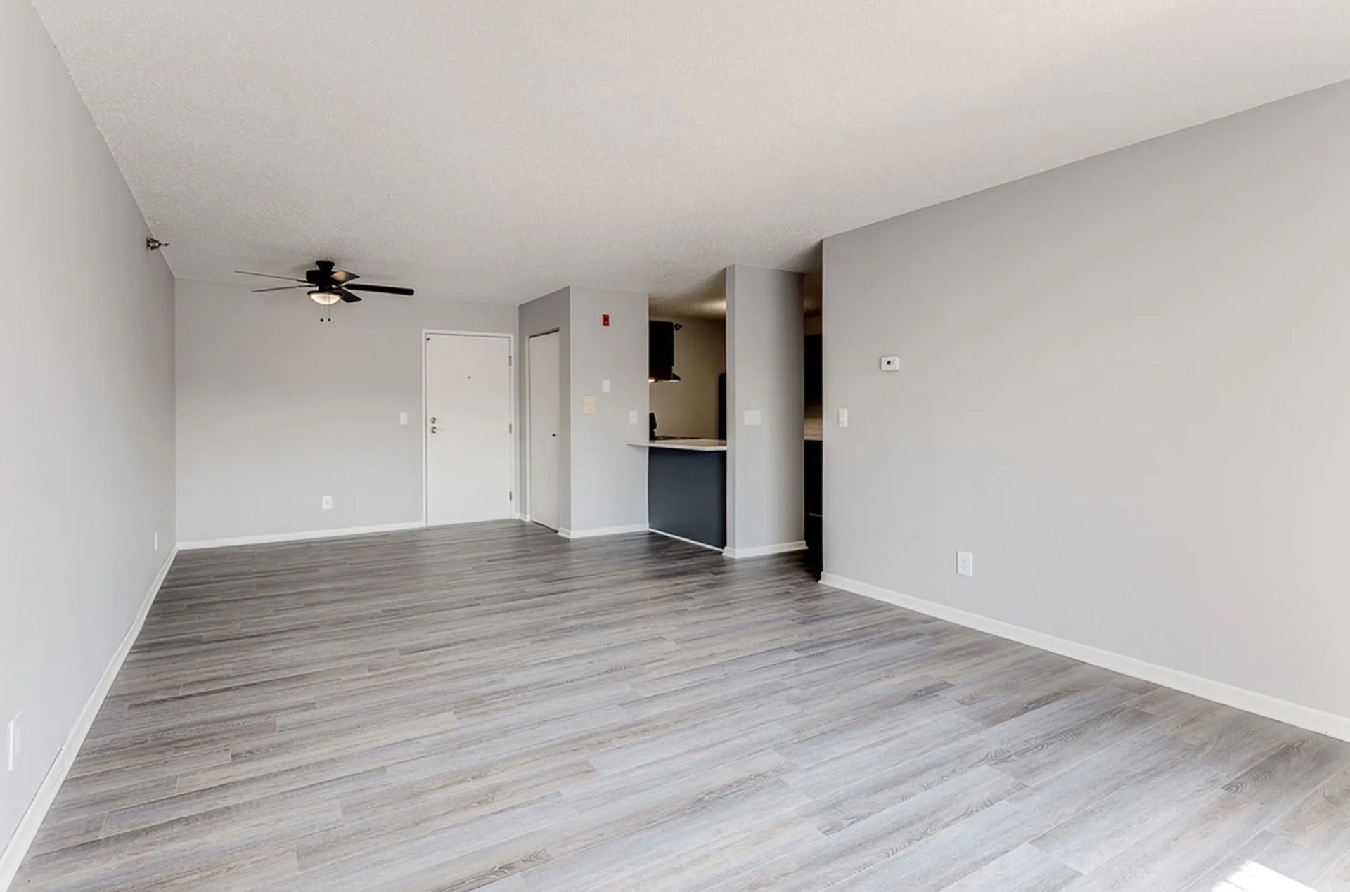 Empty living room with gray walls, wood-look flooring, and a glimpse of a kitchen.