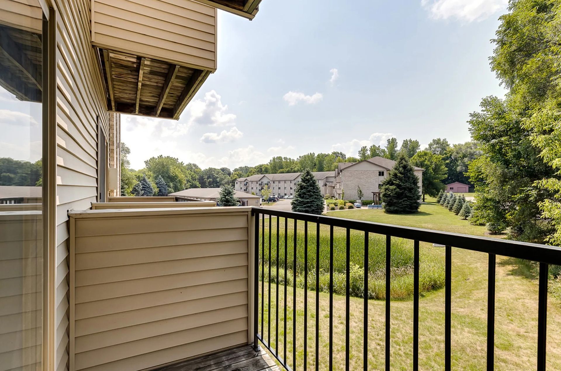 Balcony view: Black railing overlooks a grassy area with trees and buildings under a sunny sky.