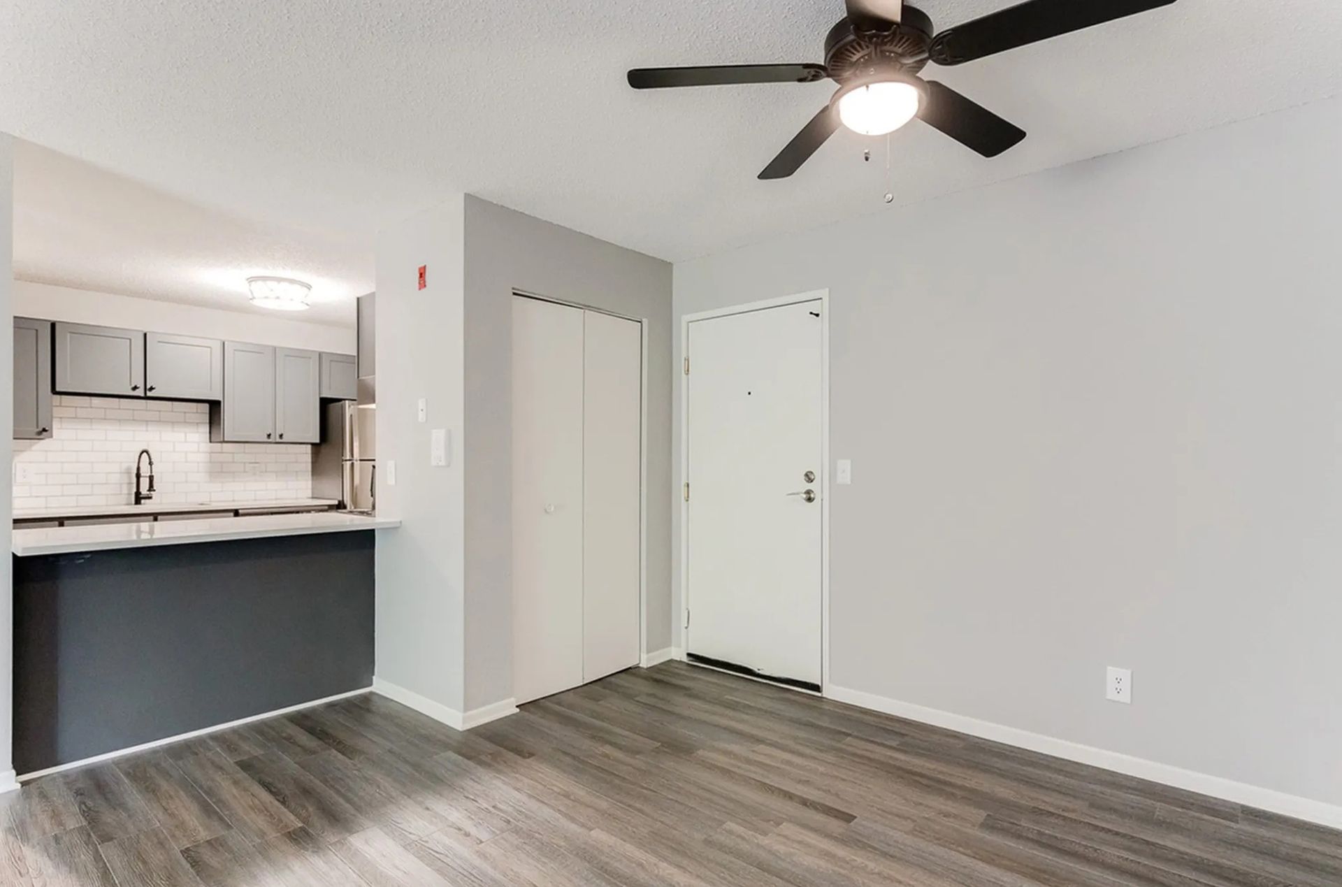 Interior of a modern apartment with kitchen, closet, front door, ceiling fan, and gray flooring.